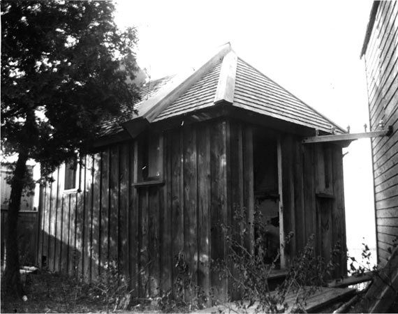Black and white photo of a small building made of vertically positioned wood planks with a shingle roof, an open door and three small windows