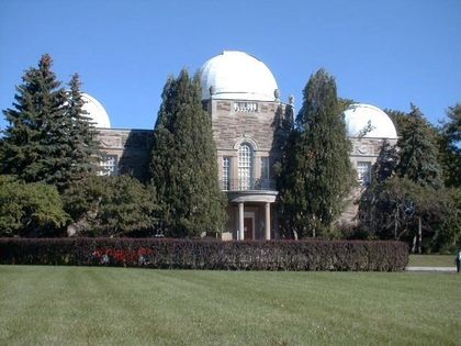 Colour photo of a grey building with three white domes, partially hidden by tall coniferous trees.