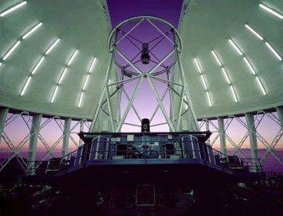 Photo taken inside an observatory where the base of the walls are raised, the white dome is open and a telescope is pointing towards a purple sky at dusk