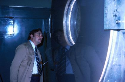 Photo d'un homme portant un manteau beige et une cravate regardant dans la fenêtre circulaire d'un mur métallique