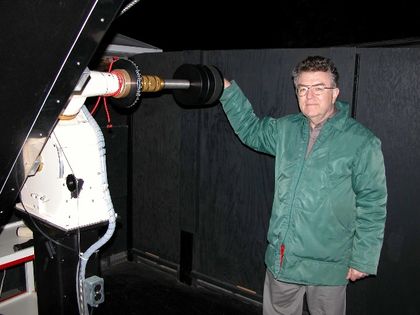Photo d'un homme portant des lunettes et un manteau vert, appuyé sur un mécanisme métallique