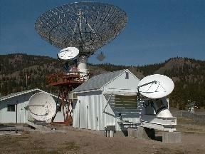 Photo couleur d'un radiotélescope grillé entouré de trois antennes blanches pointant toutes vers le ciel, autour de deux bâtiments gris avec des montagnes boisées à l'arrière.