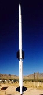 Photo couleur d'une fusée blanche, avec des ailerons noirs installés en position de décollage sur un sol sablonneux avec des montagnes à l'horizon.