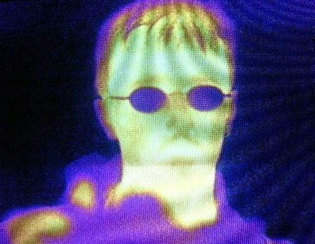 Infrared image of a human head with green, yellowish purple and bluish tinges. The person is wearing glasses easily distinguished on the face owing to their blue colour.