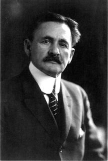 Photo en noir et blanc d'un homme portant une moustache et un complet noir.