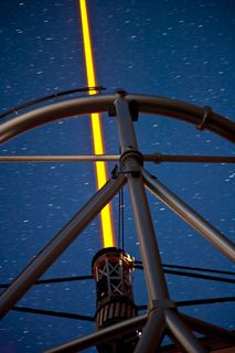 Photo d'une structure en acier avec, au centre, un faisceau jaune pointant vers le ciel étoilé la nuit.