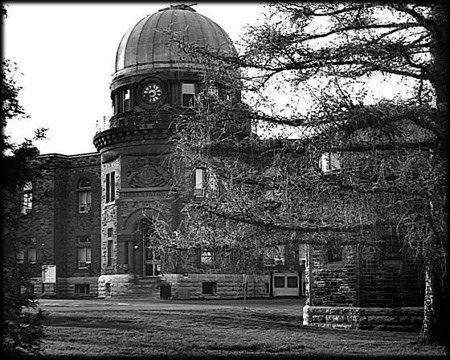 Black and white photo of a stone building with a domed roof, partially hidden by a tree.