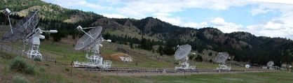 Panoramic photo of five white white antennas pointing towards the sky, located in a field with forested mountains in the background.