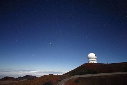 Photo d'un paysage avec à droite, un observatoire blanc, situé sur une colline brune dénudée, sous un ciel étoilé passant du blanc au bleu nuit