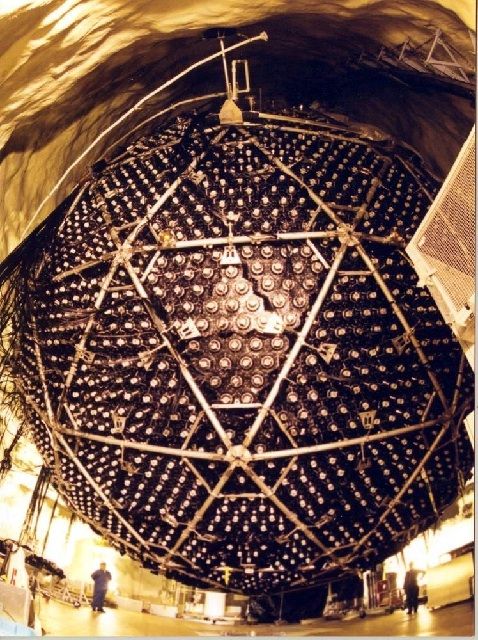 Colour photo of of a suspended brown sphere made of triangular metal components. Men are standing at each side of the sphere.