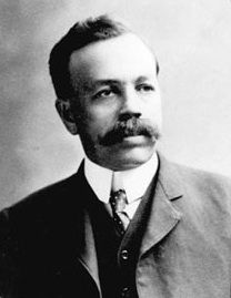 Photo en noir et blanc d'un homme regardant à droite, portant une longue moustache et un complet avec une cravate