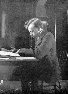 Black and white photo of a man with a mustache seated at a desk, leaning over some papers