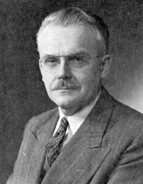 Black and white photo of a man with glasses and a mustache wearing a suit and tie