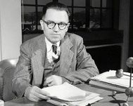 Photo noir et blanc d'un homme en complet et cravate, assis derrière un bureau, tenant des papiers dans sa main