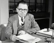 Black and white photo of a man wearing a suit and tie, seated at a desk and holding papers in his hand