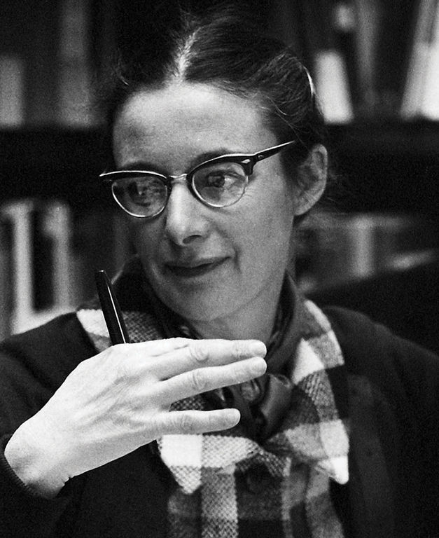Black and white photo of a woman with glasses gesticulating in front of her shoulder with her right hand