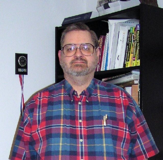 Colour photo of a man with glasses and a beard, wearing a checkered shirt, taken in front of a black bookcase and a white wall
