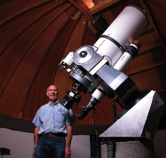 Low-angle photo of a man standing in front of a white telescope with a wooden dome in the background