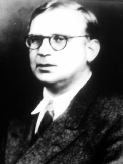 Photo en noir et blanc un peu floue d'un homme portant des lunettes rondes et un veston noir