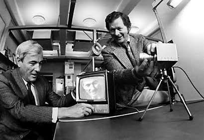 Photo of of a man filming another man using a camera installed on a table and displaying the image directly on a television screen