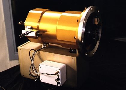 Photo d'une caméra, composée d'une structure métallique dorée de forme tubulaire raccordée par des fils et installée sur base de bois rectangulaire