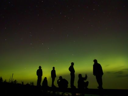 Photo of silhouettes of people standing on a hill looking at green Northern Lights in a black sky